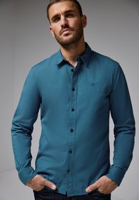 Tyrkysová košile s dlouhým rukávem na knoflíky s čistým designem, černými knoflíky a detailem loga na hrudi, v kombinaci s tmavými džíny.