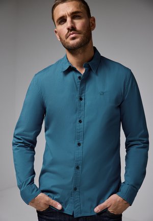 Camisa de botones de manga larga en tono azul verdoso con un diseño limpio, botones negros y un detalle de logo en el pecho, combinada con jeans oscuros.