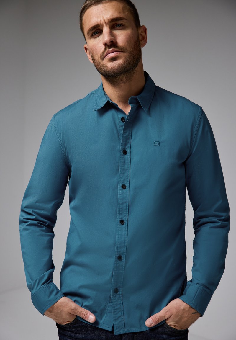 Tyrkysová košile s dlouhým rukávem na knoflíky s čistým designem, černými knoflíky a detailem loga na hrudi, v kombinaci s tmavými džíny.