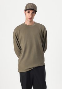 Cămașă lungă cu mânecă, de culoare verde măsliniu și cu un guler rotund, purtată cu pantaloni negri; purtând o șapcă de baseball în carouri cu bor întunecat.