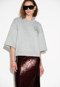 Vrouw in een oversized grijze sweatshirt met geborduurd logo, een donkere sequined rok, grote zwarte zonnebril, zilveren armband en oor cuff.