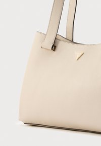 Beżowa skórzana torba typu tote o fakturowanej powierzchni, z wydłużonymi paskami i trójkątnym złotym elementem ozdobnym z nazwą marki.