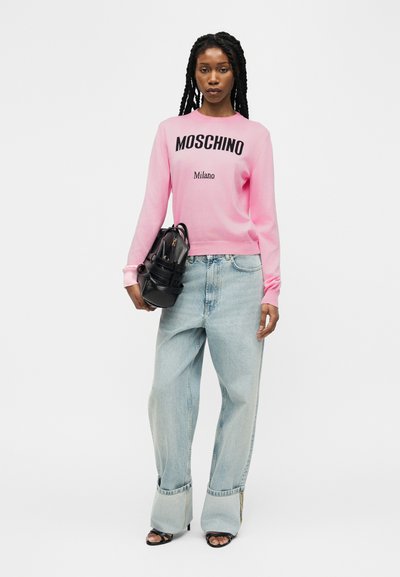 Femme portant un pull Moschino Milano rose, un jean pattes d'éléphant bleu clair, des sandales noires, et tenant un sac à main noir.