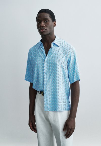 Jeune homme portant une chemise à manches courtes bleu clair à motifs et un pantalon blanc, debout devant un fond gris uni.