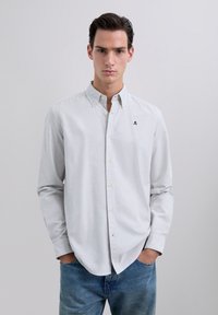 Giovane uomo che indossa una camicia a righe sottili grigia chiara con un piccolo logo a teschio, jeans blu, mani in tasca, sfondo neutro.