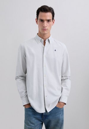 Jeune homme portant une chemise boutonnée à rayures grises claires avec un petit logo de skull, un jean bleu, les mains dans les poches, fond neutre.