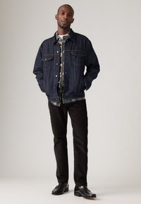 Chaqueta de denim en azul oscuro, con cierre de botones, dos bolsillos en el pecho, superpuesta sobre una camisa de cuadros. Pantalones de pana negra y zapatos negros completan el conjunto.