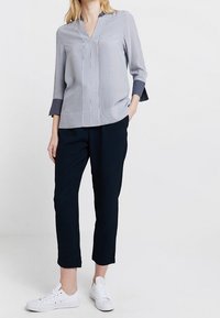 Žena v světle modré dlouhé haleně s pruhovanými manžetami, černých zkrácených kalhotách a bílých teniskách, stojící s jednou rukou v kapse.