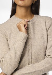 Beige strikket cardigan med rund hals, ribbestrikkede mansjetter og fem knapper foran. Myk tekstur med et subtilt mønster.