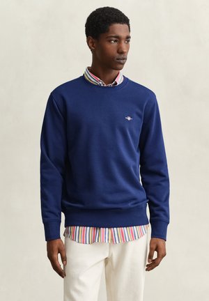 Junger Mann, der ein blaues Sweatshirt über einem mehrfarbigen gestreiften Hemd und weißen Hosen trägt, vor einem schlichten beigen Hintergrund steht.