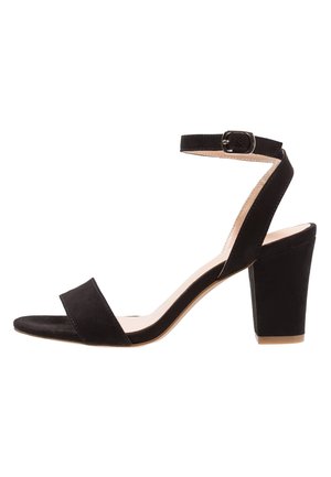 Sandale pour femme en daim noir avec talon bloc, bout ouvert et bride à la cheville avec boucle argentée.