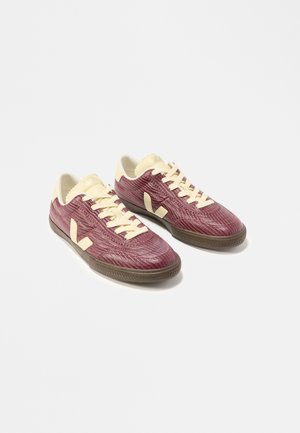 Paire de baskets basses avec tiges bordeaux texturées, lacets crème, languettes et accents au talon, et semelles en caoutchouc marron sur fond blanc.