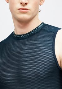 Detailný záber osoby nosiacej čierny sieťovaný top bez rukávov s textom "LE COL" opakujúcim sa na golieri.