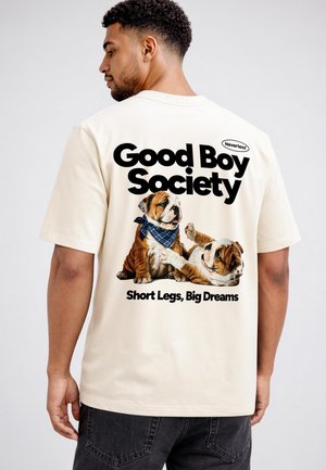 Mann trägt ein weißes T-Shirt mit zwei verspielten Bulldog-Welpen und den Texten "Good Boy Society" und "Short Legs, Big Dreams" auf dem Rücken gedruckt.