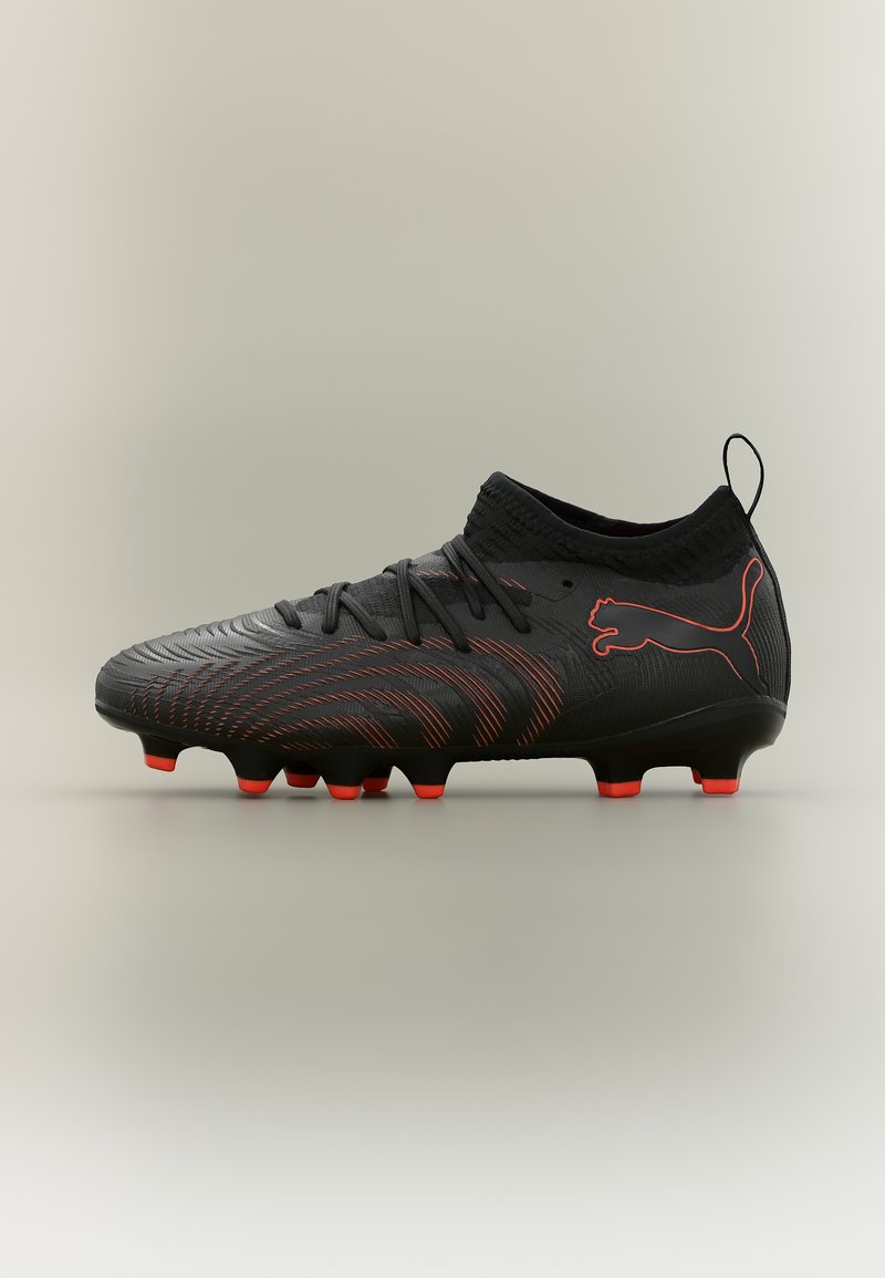 Μαύρο και κόκκινο παπούτσι ποδοσφαίρου Puma με κορδόνια, υφή στην επιφάνεια, ψηλός γιακάς αστραγάλου και καρφιά στη σόλα, πάνω σε μπεζ φόντο.