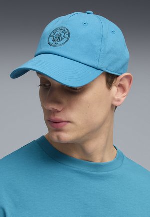 Młody mężczyzna w jasnoniebieskiej czapce Manchester City i pasującej niebieskiej koszuli, spogląda w dół i na bok na tle szarego tła.