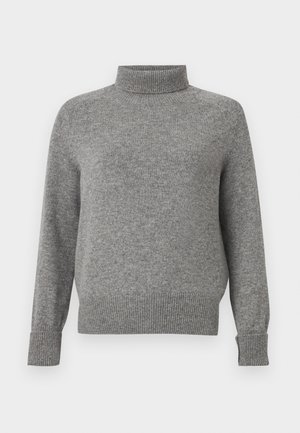 Kobieta ubrana w szary sweter z golfem, czarne szerokie spodnie oraz czarne, odkryte na palce szpilki, stojąca na białym tle.