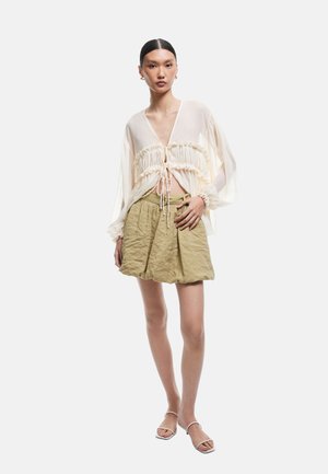 Lightweight, sheer cream blouse with elastic ruffles at the waist, paired with a textured olive green mini skirt and simple strappy sandals.
