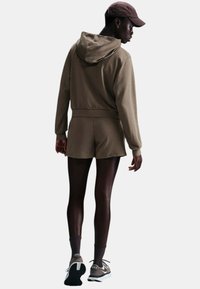 Felpa con cappuccio marrone abbinata a pantaloni corti dello stesso colore, indossata con collant scuri e sneakers, vista da dietro. Design minimalista, tessuto morbido.