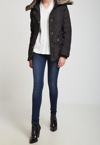 Svart vadderad pufferjacka med huva i fuskpäls, kombinerad med en vit plisserad blus, mörka skinny jeans och glänsande svarta ankle boots.