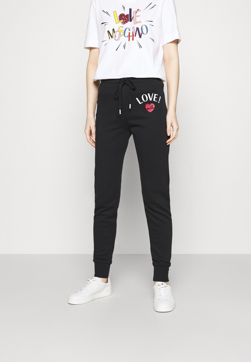 Black sweatpants with 'LOVE!' and a heart design in red glitter, featuring an elastic waistband and cuffs. Paired with a white graphic tee.