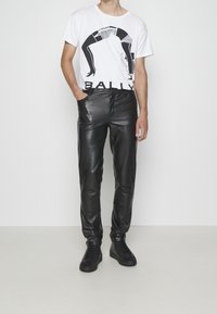 Vit T-shirt med svart grafisk design och "BALLY"-text, kombinerad med åtsittande svarta läderbyxor och svarta hög-vrist sneakers.