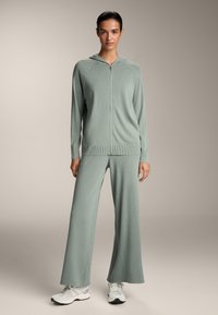 Svijetlozeleni rebrasti komplet za opuštanje koji se sastoji od zip-up dukserice i širokih hlača, izrađen od mekog materijala s opuštenim krojem i minimalističkim dizajnom.