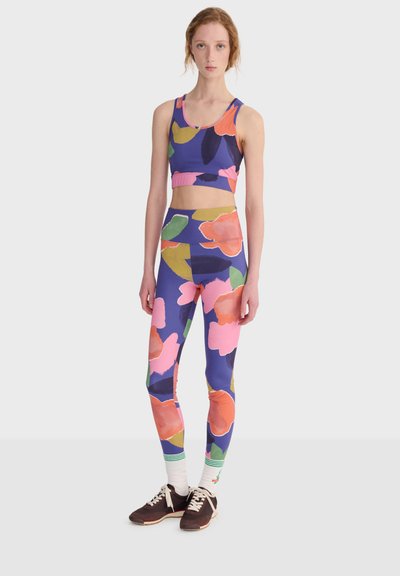 Femme debout portant un brassière de sport bleu à fleurs et des leggings, des chaussettes blanches avec des rayures vertes, et des baskets marron foncé sur un fond uni.