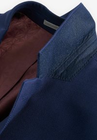 Granatowa kurtka z teksturowanej tkaniny z gładką bordową podszewką. Wyróżniające się szczegóły to usztywniony kołnierz i staranne szwy.