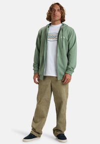 Une hoodie zippée vert menthe, un t-shirt blanc avec un logo bleu et vert, un pantalon en velours côtelé beige et des chaussures navy. Style décontracté, coupe relax avec poches.