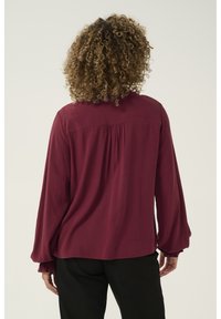 Maroon blouse with long, puffy sleeves and ruffled cuffs. Back features a gathered seam for added detail. Soft fabric drapes gently.