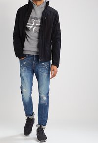 Chaqueta negra con bolsillos con cremallera sobre una sudadera gris, combinada con jeans azules desgastados y zapatillas negras. Atuendo casual en capas.