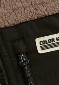 Tissu en polaire marron au-dessus d'un tissu noir avec fermeture éclair et tirette en cordon noir et blanc ; étiquette partielle "COLOR K" sur le tissu noir.