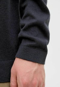 Mørkeblå strikkegenser i nærbilde, som viser ribbestrikket mansjett, jevn tekstur og en tettsittende fasong med en hånd som griper om ermet.