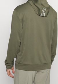 Sweat à capuche vert olive avec une texture lisse, doté d'une capuche à cordon et de détails subtils du logo au dos. Manches longues et coupe décontractée.