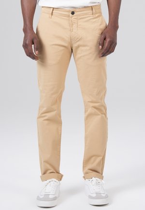 Chinos beige à coupe slim, confectionnés en coton, avec une fermeture à bouton, des poches latérales et des poignets roulés, associés à des baskets blanches.