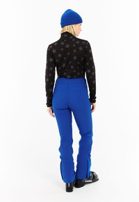 Zwarte top met lange mouwen en sterrenpatroon, gecombineerd met blauwe skibroek, zijritsen en zwarte skischoenen. Model draagt een blauwe muts.