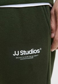 Olivově zelený materiál joggerů má ribovanou strukturu, bílou značkovou logo "JJ Studios" s dalším textem na boku a přiléhavý design.