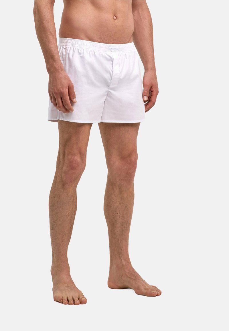 Boxer en coton blanc avec une ceinture élastique, présentant un design simple et une coupe décontractée, porté par un modèle debout pieds nus.