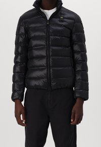 Man in een zwart glanzend pufferjack, dichtgeritst, gecombineerd met zwarte broek en een wit shirt eronder.