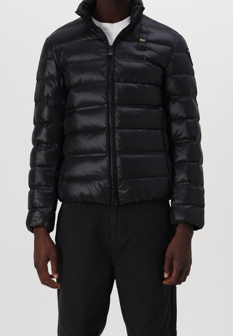 Man in een zwart glanzend pufferjack, dichtgeritst, gecombineerd met zwarte broek en een wit shirt eronder.