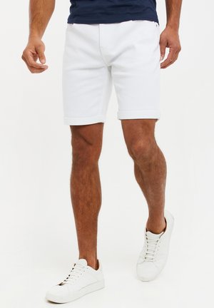 Homme marchant portant un short blanc retroussé, des baskets blanches et une chemise bleu marine sur fond blanc.