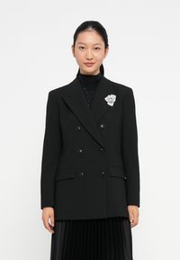 Blazer nero doppiopetto con rever a scalfo, due tasche frontali e un accento in spilla floreale argento. Texture liscia, design strutturato.