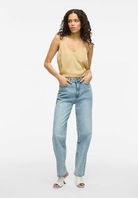Top de satén beige sin mangas con tirantes finos combinado con jeans de tiro alto en azul claro. La modelo lleva sandalias blancas de tacón abierto. Fondo limpio.