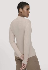 Beige geripptes Langarmoberteil mit hohem Kragen und figurnaher Silhouette, kombiniert mit braunen Hosen, die aus strukturiertem Stoff bestehen und schmale Ärmel haben.