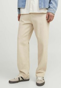 Beige volné kalhoty vyrobené z bavlny s hladkou texturou. V páru se šedými teniskami s černými akcenty a bílými tkaničkami.