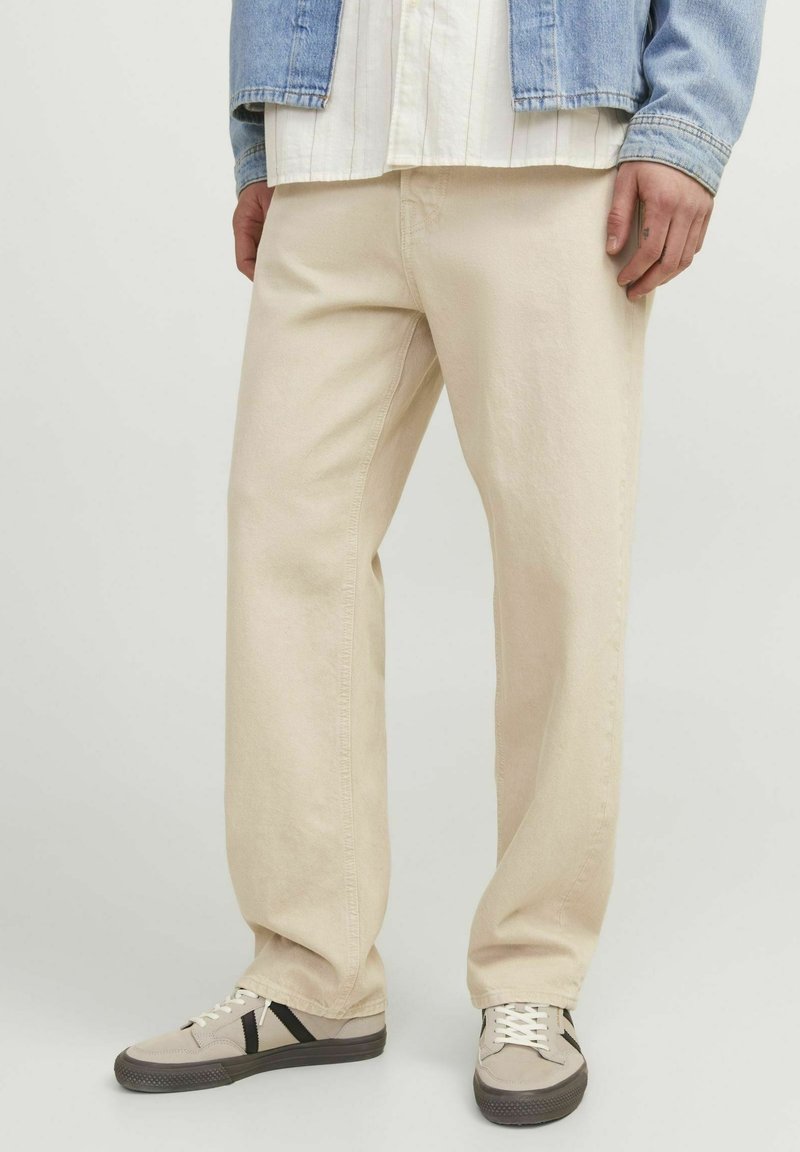 Beige volné kalhoty vyrobené z bavlny s hladkou texturou. V páru se šedými teniskami s černými akcenty a bílými tkaničkami.