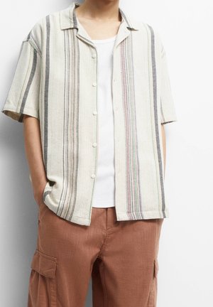 Mand iført kortærmet skjorte med knapper og lodrette beige, grå og pastelfarvede striber over en hvid tanktop og rustfarvede cargobukser.