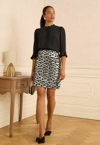Donna che indossa una blusa nera e una gonna a motivi argentati, con una borsa tonda in mano, in piedi accanto a un tavolo di legno con una lampada d'oro.