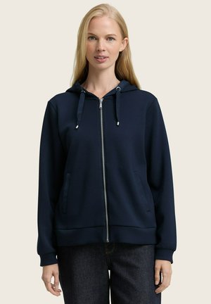 Femme blonde portant un hoodie zippé bleu marine avec des cordons, se tenant avec les mains détendues sur les côtés contre un fond uni.